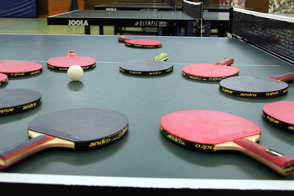 Mehrere Tischtennisschläger in verschiedenen Farben und ein Tischtennisball liegen auf einer grünen Tischtennisplatte.