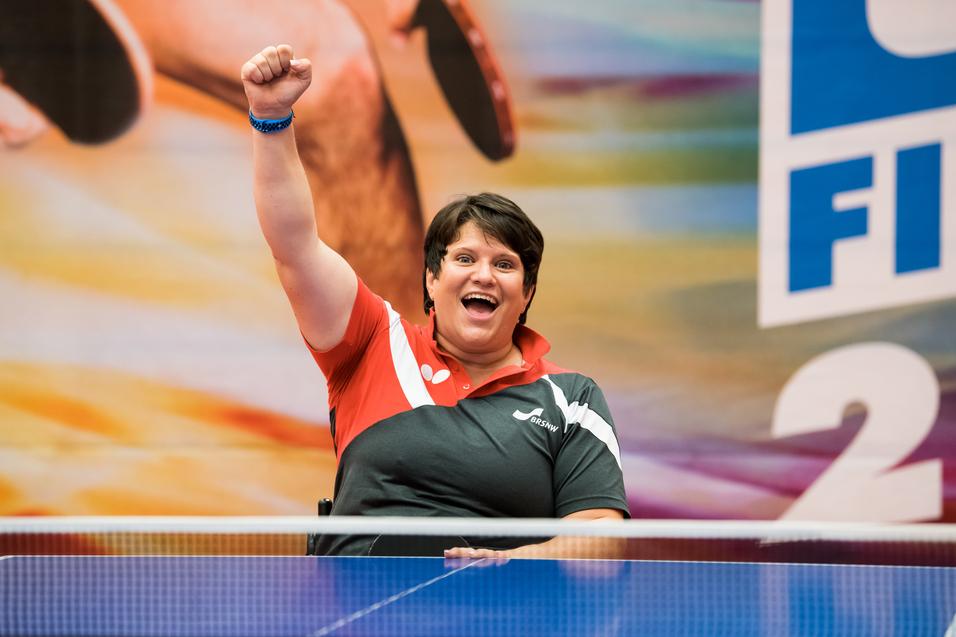 Eine jubelnde Frau mit kurzen Haaren in einem rot-schwarzen Sportoutfit erhebt den Arm vor einem bunten Hintergrund.